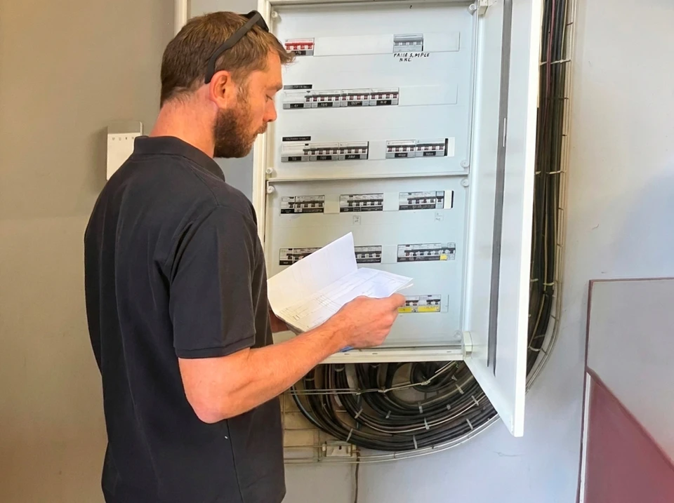 Technicien en polo gris consulte des plans papier devant un tableau électrique industriel ouvert